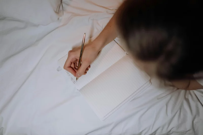 A person writing a to-do list in a notebook while sitting on a bed with white linen.