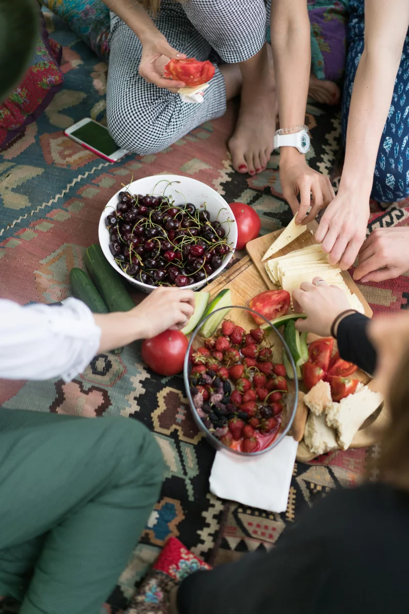 Casual gathering with hands reaching for cherries, strawberries, and bread on a colorful rug.