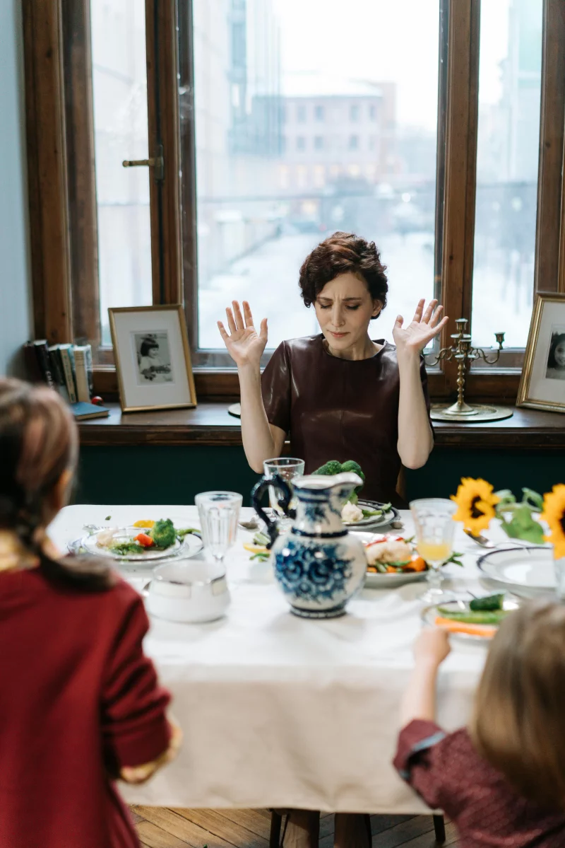A mother appearing stressed at the dinner table with her two children indoors.