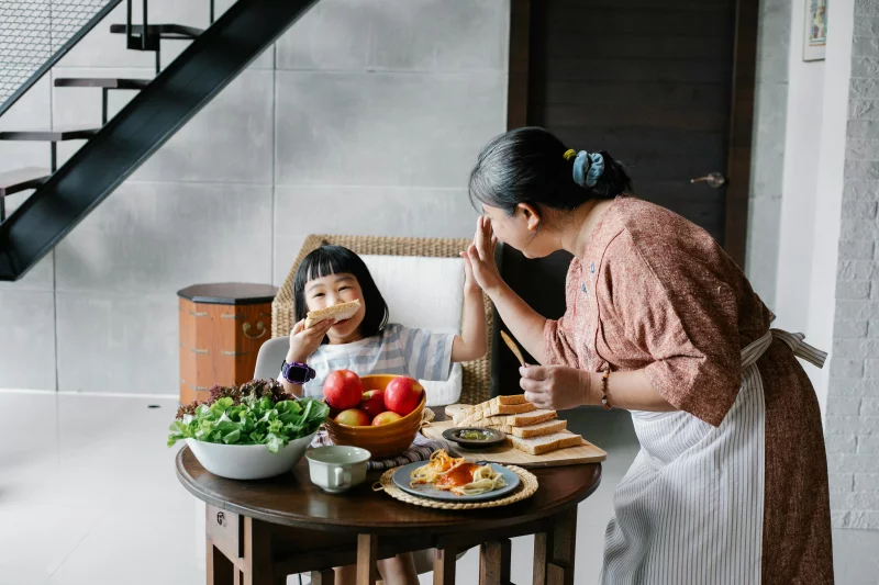Asian grandmother and granddaughter sharing a joyful breakfast with toast and fruits indoors.