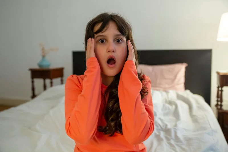 Young girl looks shocked while sitting on a bed in a cozy bedroom.
