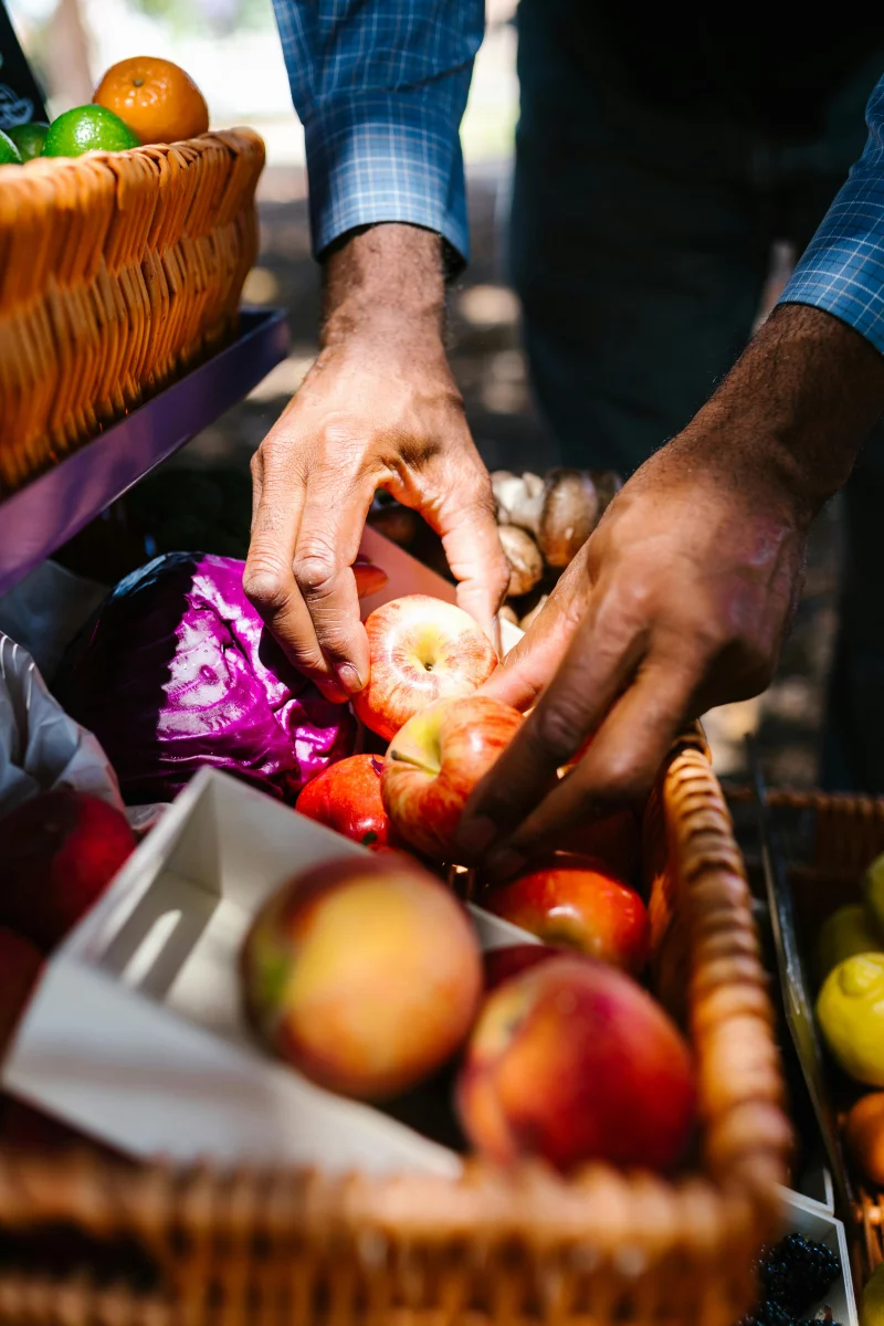 A close-up of hands selecting red apples from a wicker basket at an outdoor market.