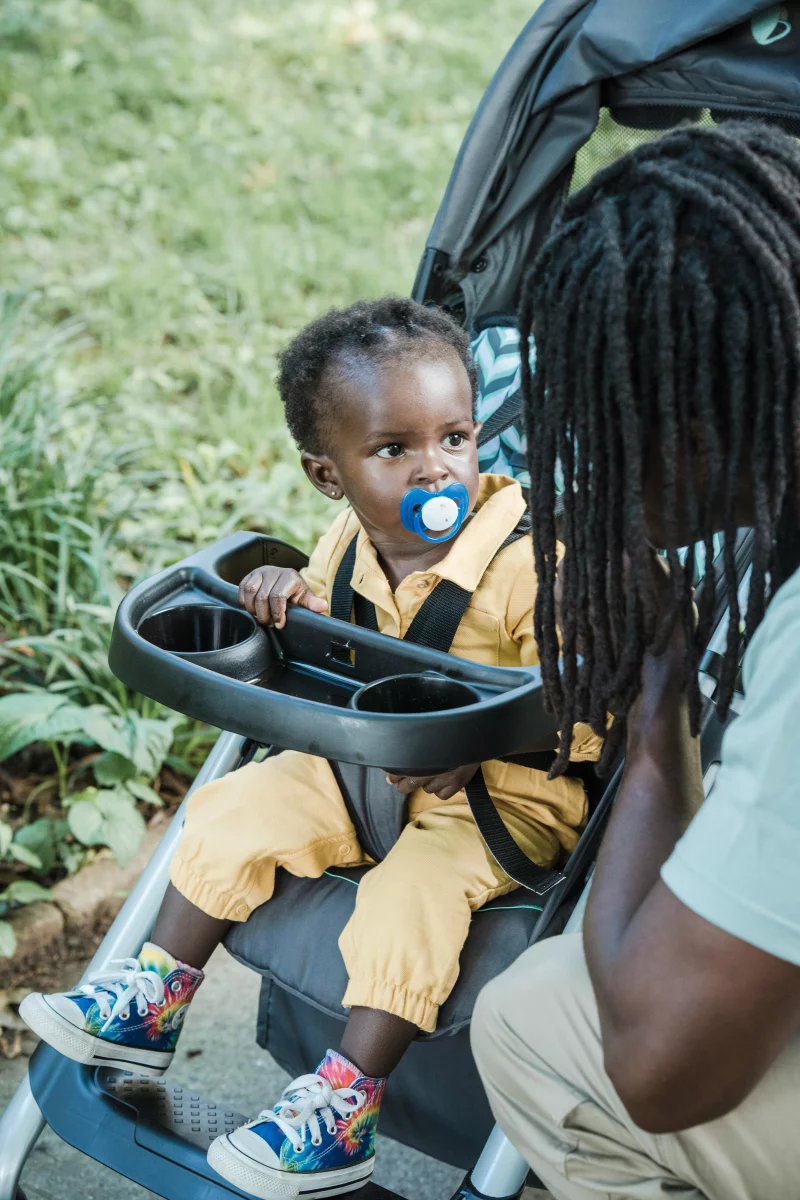 A father interacts with his toddler in a stroller during a sunny day outdoors.