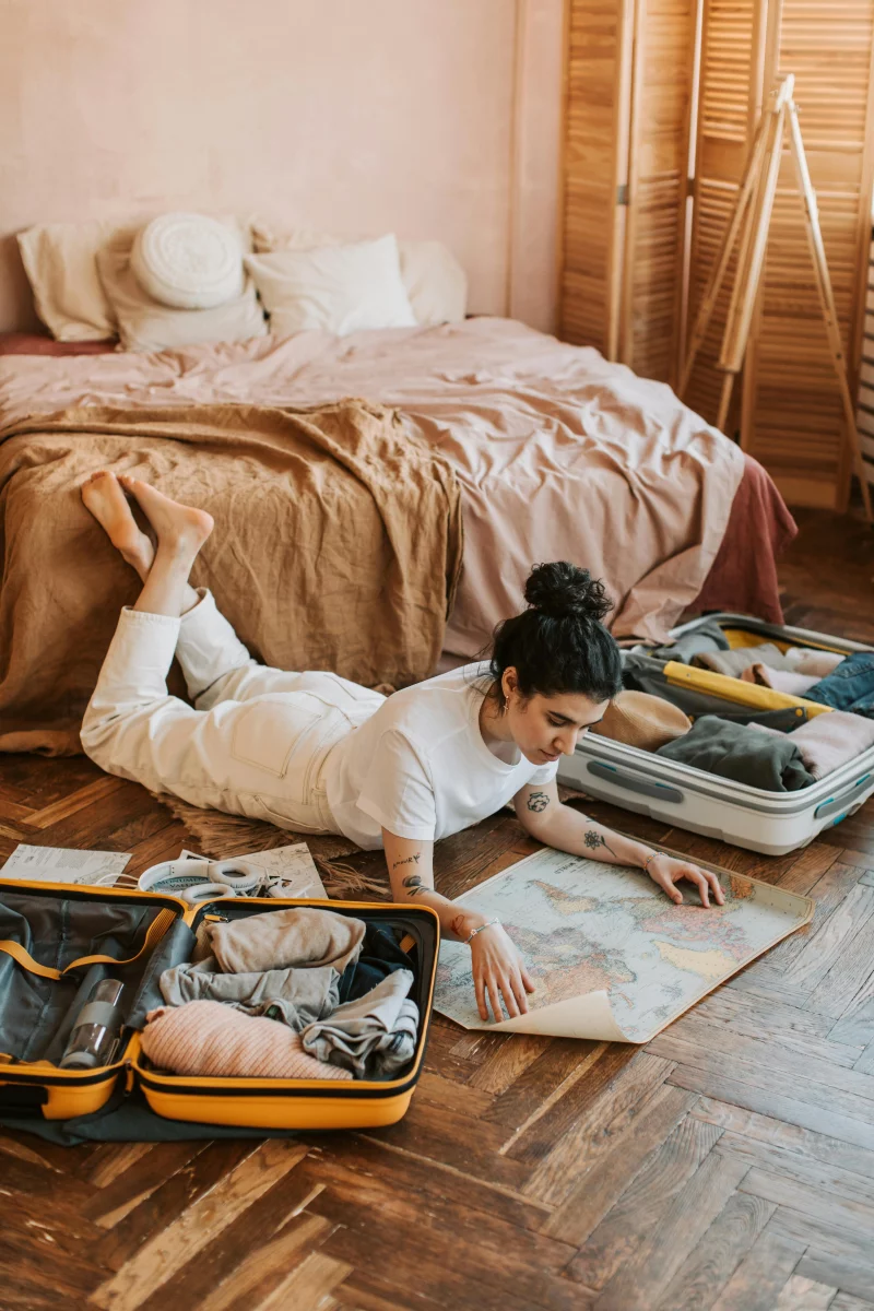 Woman lying on floor in bedroom, planning trip with map and open suitcases.