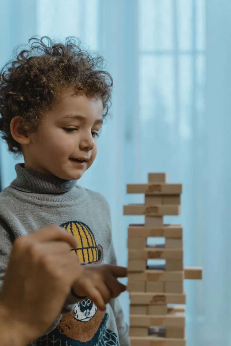 A young boy plays with wooden blocks, guided by an adult hand indoors.