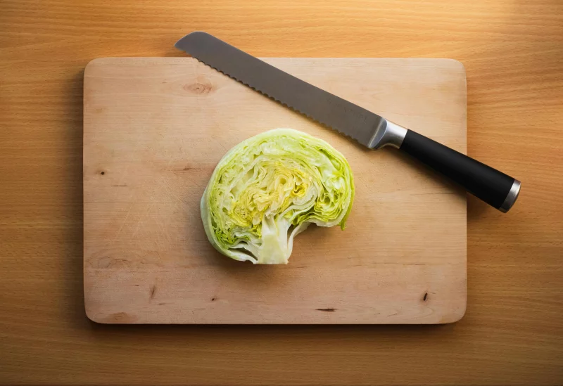Half a crisp iceberg lettuce on a wooden board with a serrated knife.
