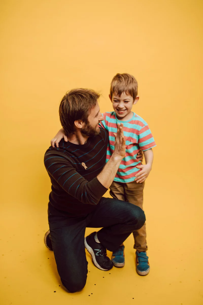Father and son sharing a joyful high five indoors against a vibrant yellow background.