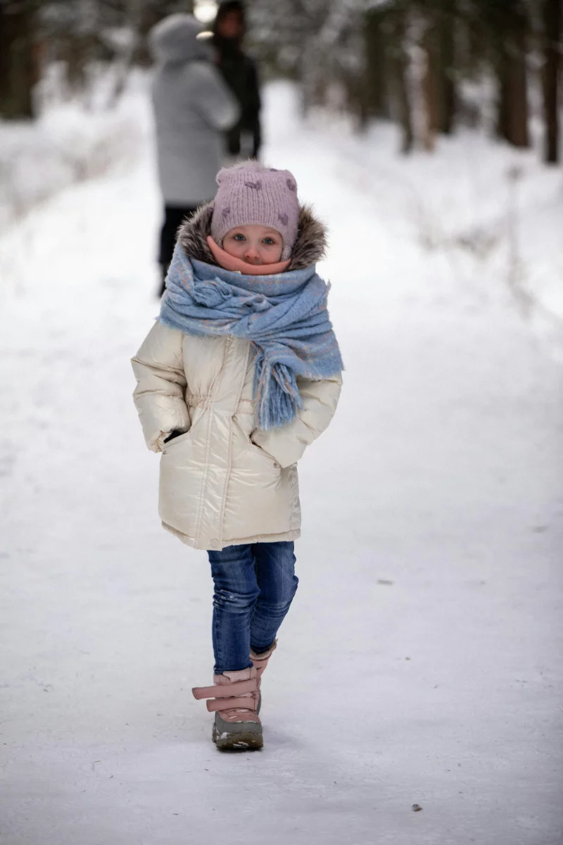 A young girl dressed for winter walks along a snowy forest path, bundled up warmly.