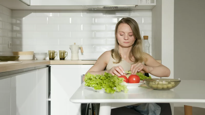 Woman sitting at table preparing fresh vegetables in a modern kitchen, highlighting healthy living and independence.