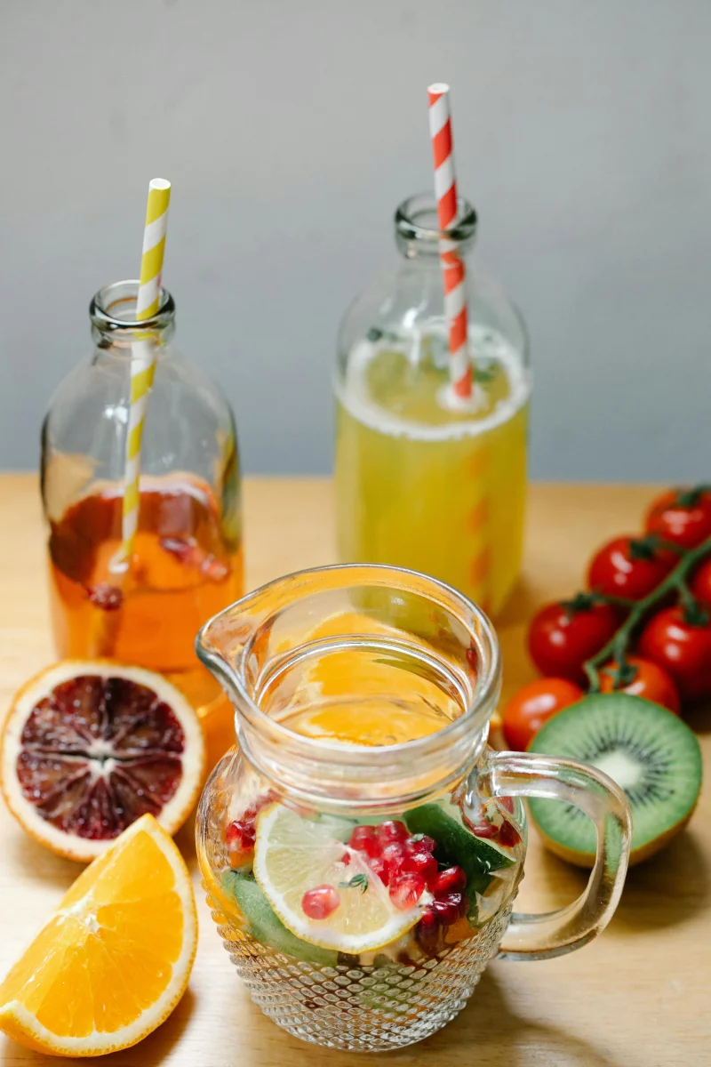 Vibrant glass pitcher and bottles with fruit-infused drinks, featuring kiwi, orange, and pomegranate.