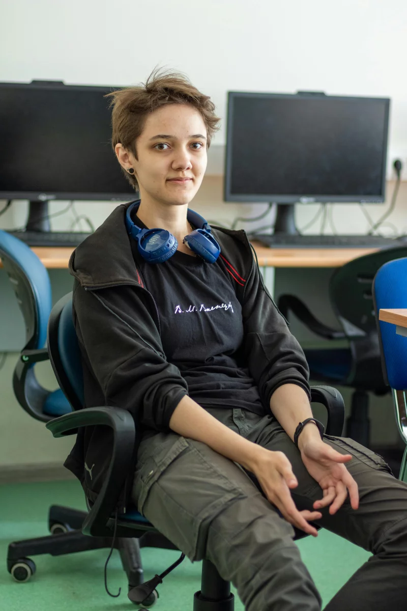 Teen sitting in a classroom wearing headphones, with computers in the background.