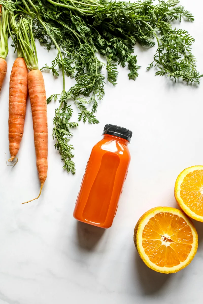 Bright and healthy carrot and orange juice bottle on white surface with fresh ingredients.