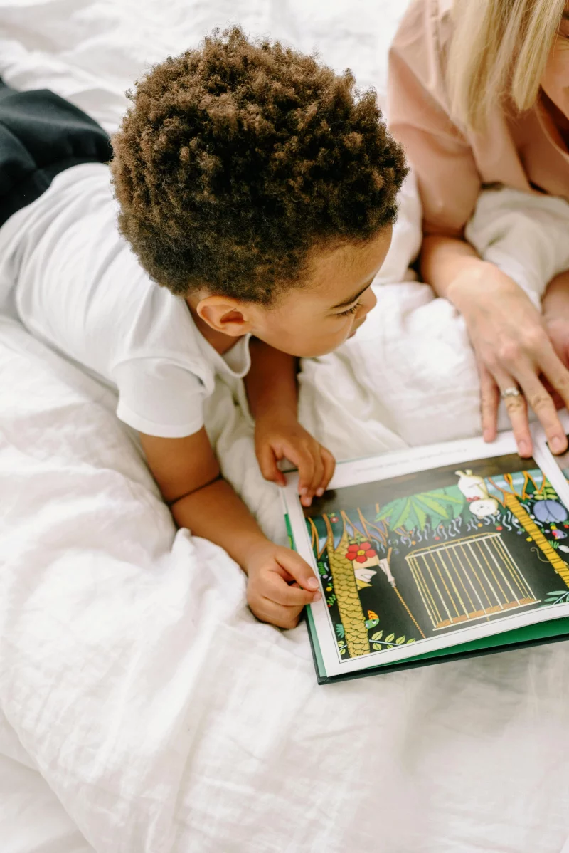 Young boy engaged in reading a picture book indoors with a parent providing guidance.
