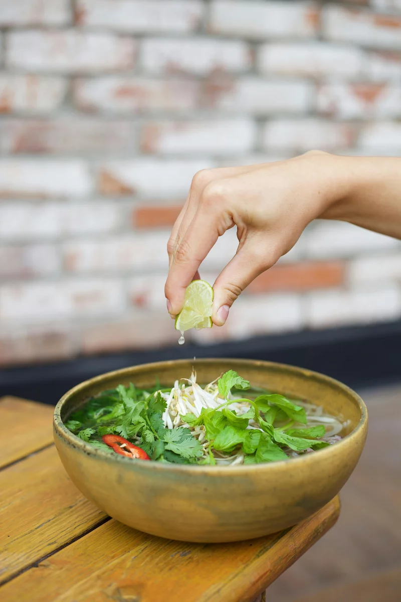 Hand squeezing lime over vibrant Vietnamese pho with herbs and chicken.