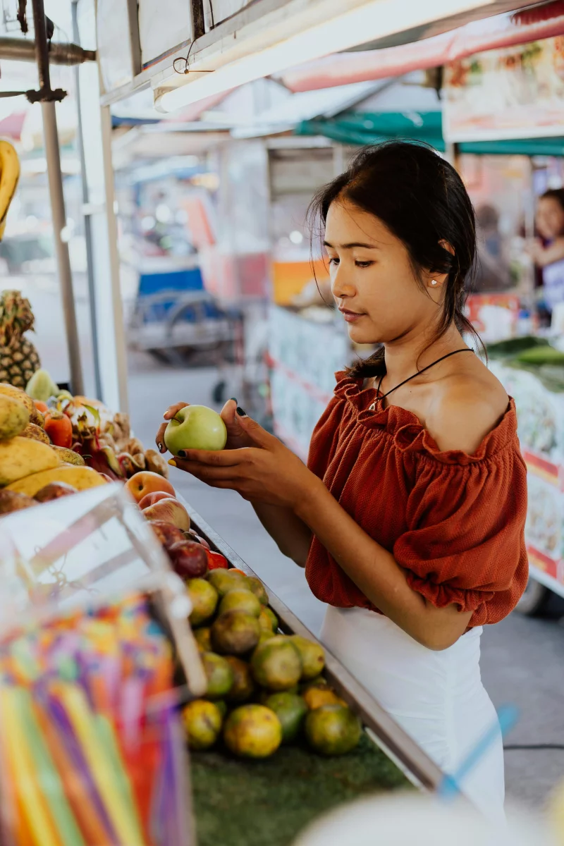 Young woman shopping for fresh fruits at an outdoor market, holding a green apple.