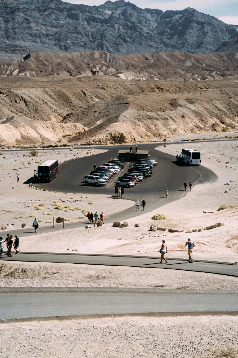 Tourists explore a desert parking area with cars and buses, set against mountain backdrop.