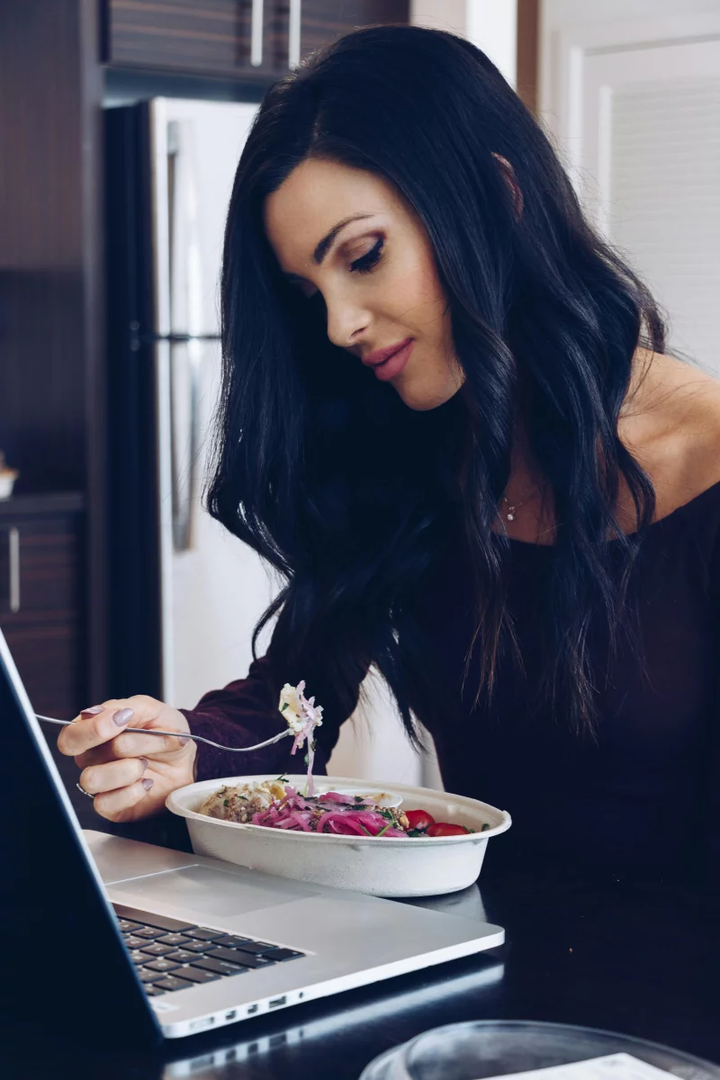 Adult woman eating a nutritious meal and working on her laptop indoors.