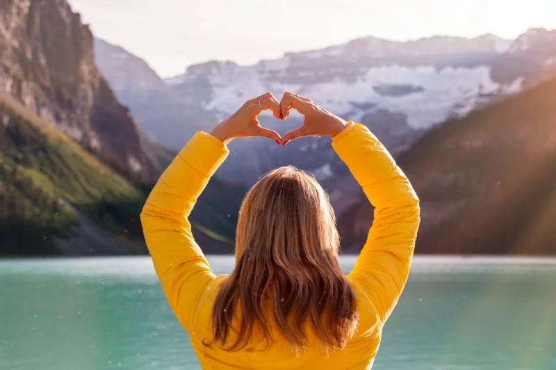 A woman in a yellow jacket forms a heart shape with her hands in front of Lake Louise's scenic backdrop.
