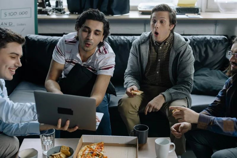 Four men in a casual office setting enjoying pizza while watching something on a laptop screen.
