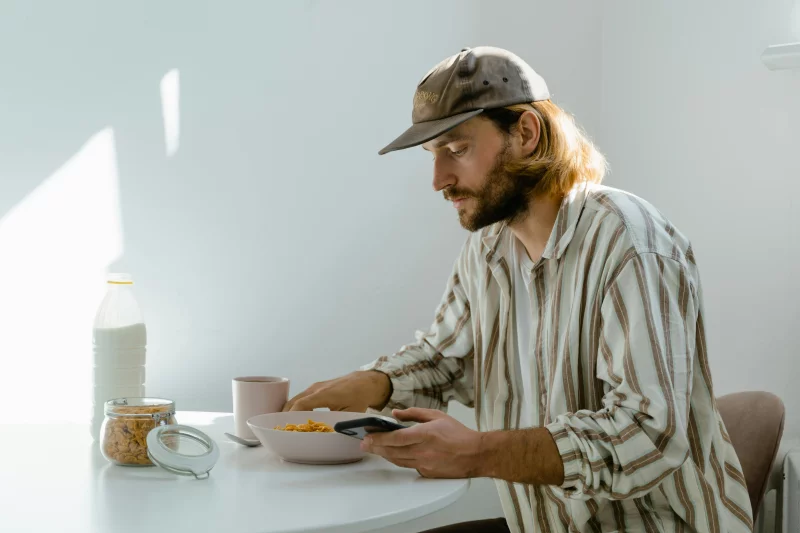 A man enjoys a casual breakfast with cereal in a sunlit room, using his phone.
