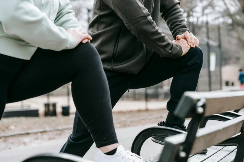 Crop faceless sportive people standing with raised legs on bench during training on street in park in daytime