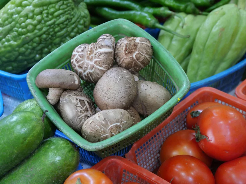 Fresh vegetables including mushrooms, bitter melon, and tomatoes in market baskets.