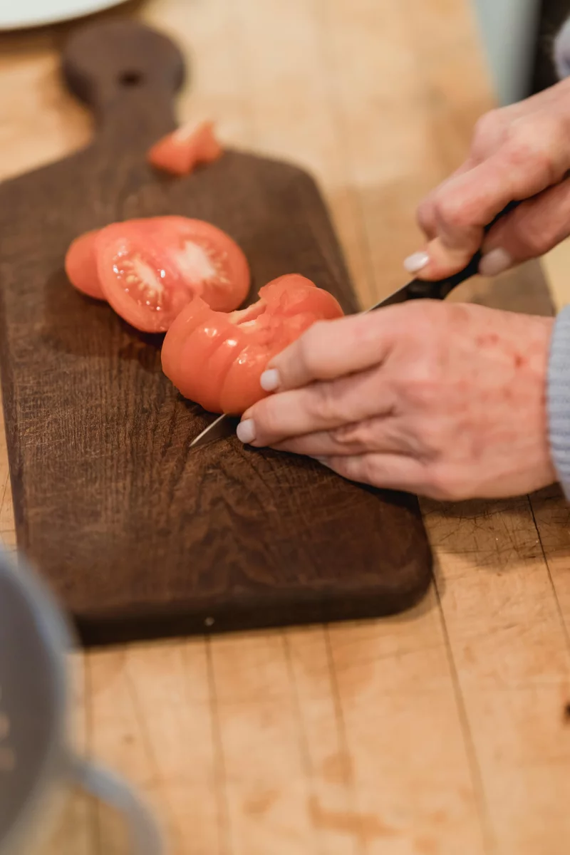 Crop unrecognizable female slicing ripe tomato on wooden board while cooking in light kitchen