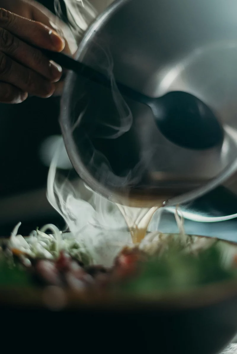 Close-up of steaming hot liquid being poured from a pot with spoon into a bowl of fresh ingredients.