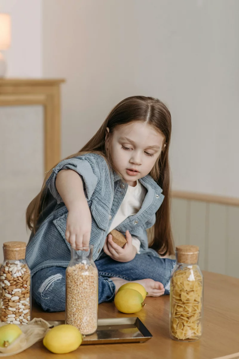 A young girl organizing jars containing pasta and grains on a wooden table in a cozy kitchen setting.