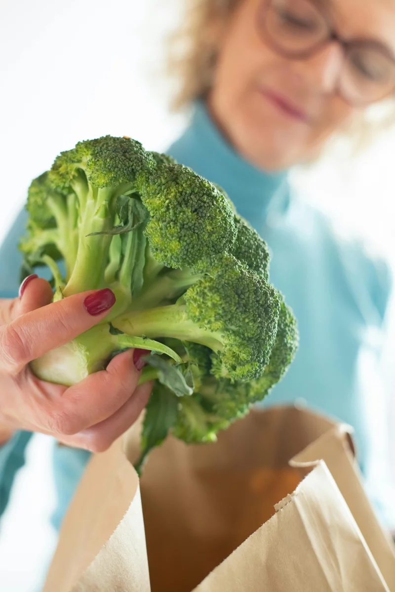A woman in a blue sweater selects fresh broccoli from a paper bag indoors.