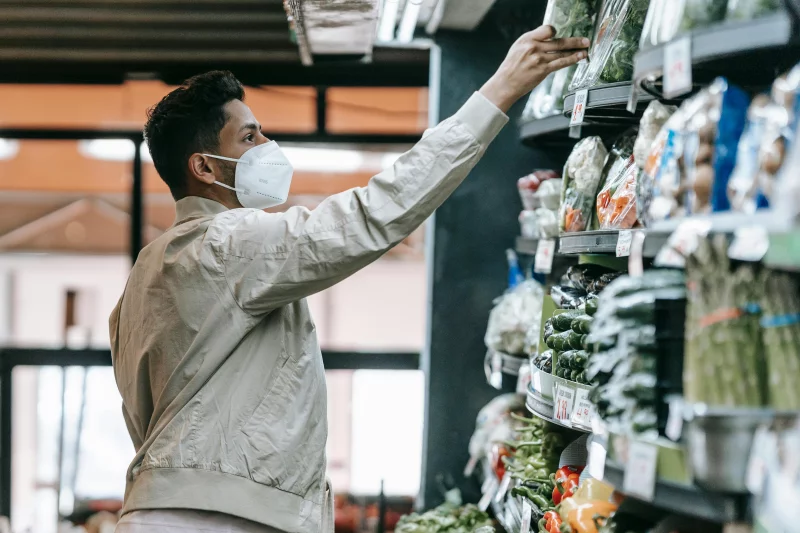 Man in a mask selecting fresh vegetables in a grocery store.