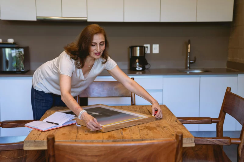 Senior woman arranging a picture frame on a wooden table in a modern kitchen setting.