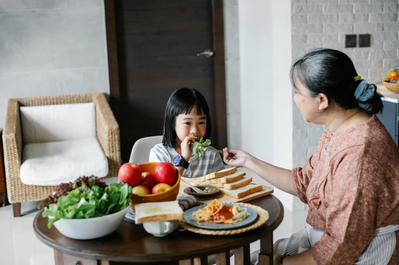 Ethnic daughter eating lettuce leaf while having healthy meal with mother in modern apartment