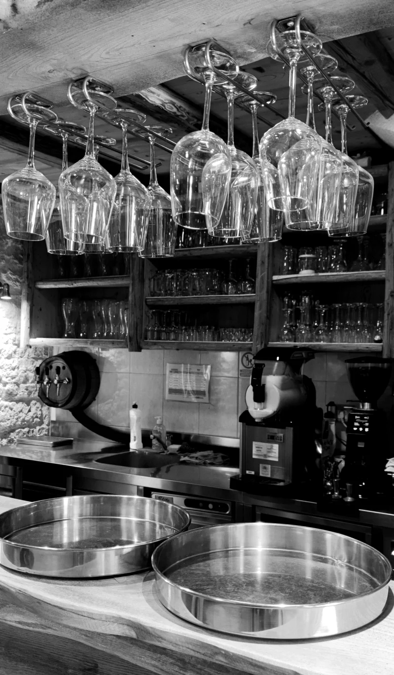 Black and white photo of a bar interior in Umag, Croatia, featuring hanging wine glasses and kitchen equipment.