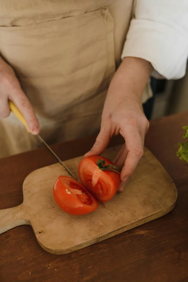 Hands slicing a fresh tomato with a knife on a wooden chopping board, showcasing food preparation.