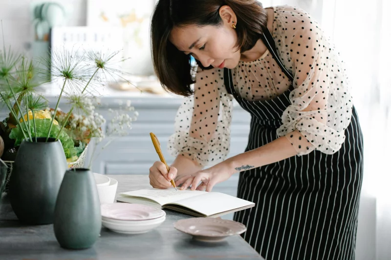 Focused woman in apron writing recipes in a cookbook in a stylish kitchen.
