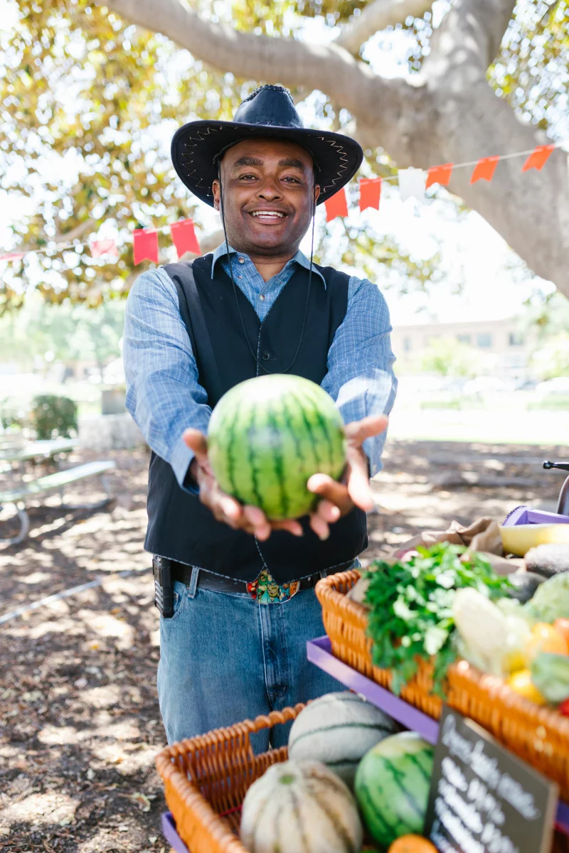 Smiling man in cowboy hat holding a watermelon at a vibrant outdoor farmer's market.