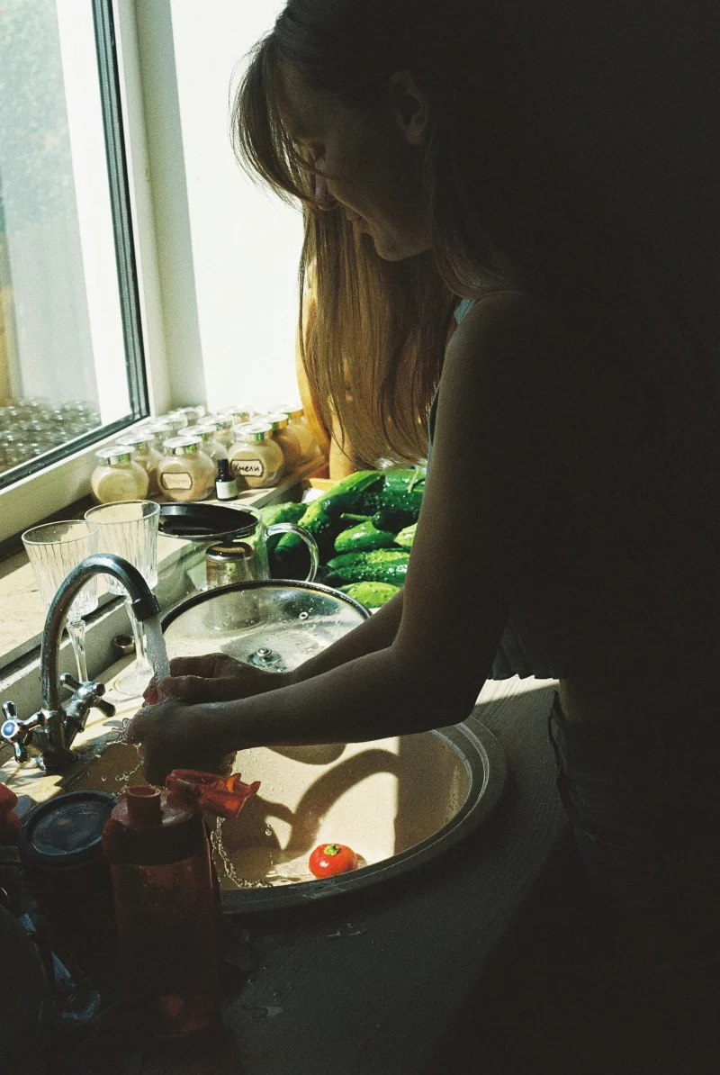 A woman washes cucumbers and tomatoes in a sunlit kitchen, creating a warm, homely atmosphere.