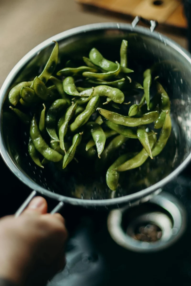 Close-up of green edamame being washed in a metal strainer, showcasing fresh food preparation.