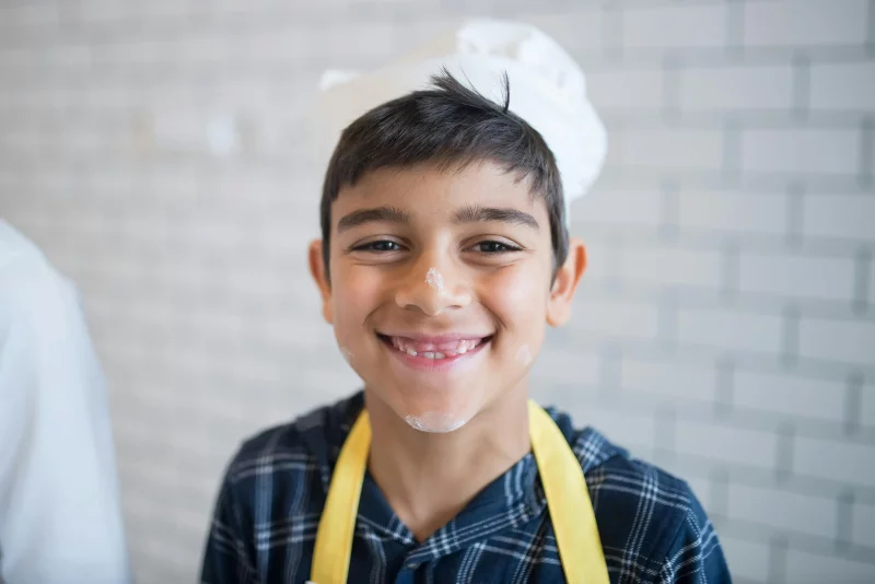 Adorable child with flour on face, wearing a chef hat and apron, smiling in a kitchen.