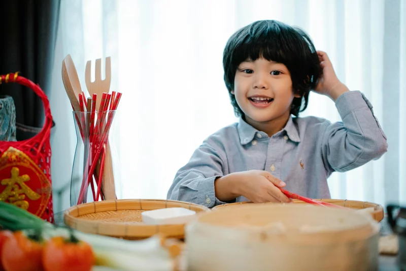 Smiling ethnic boy touching head while preparing Chinese meal at table in kitchen and looking at camera