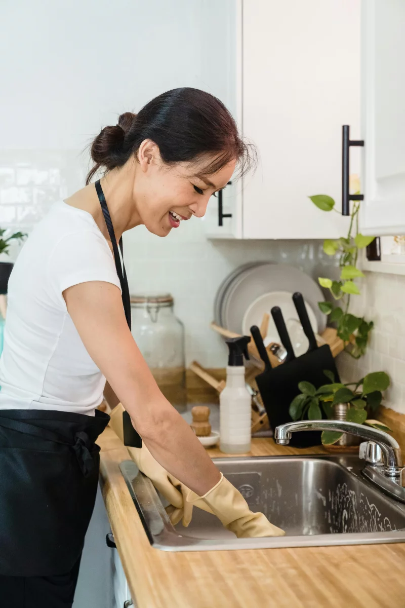 Smiling woman washing dishes with gloves in a stylish kitchen sink area.