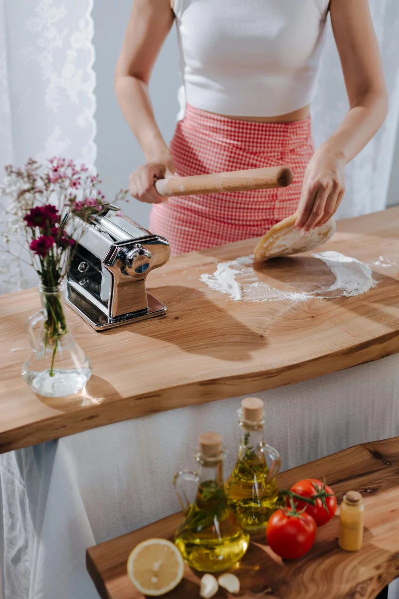 A woman preparing Italian pasta using a rolling pin and fresh ingredients in a cozy kitchen setting.