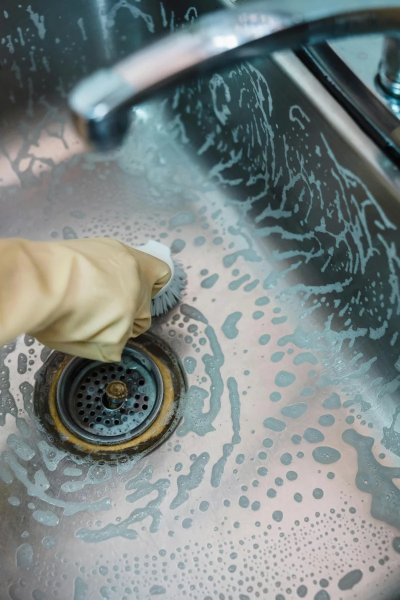 Close-up of a hand cleaning a soapy stainless steel sink using a brush.
