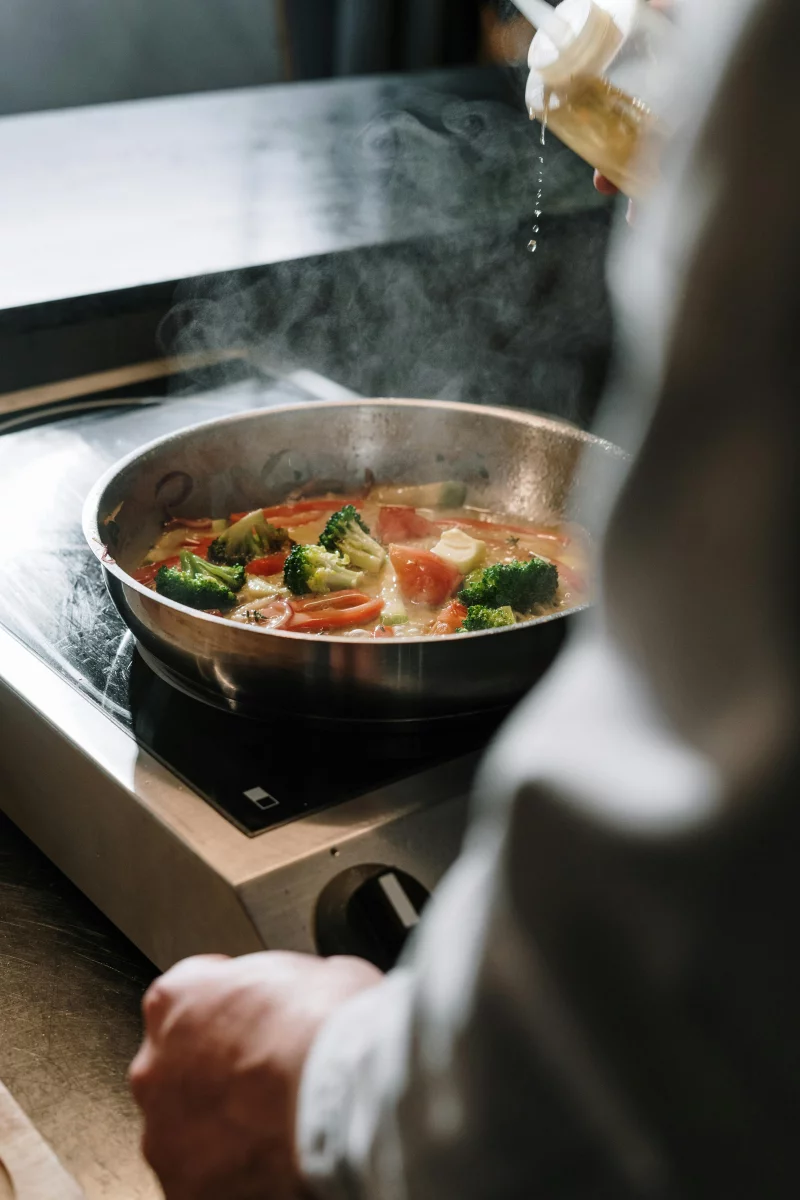 A chef is drizzling oil over steaming vegetables in a frying pan indoors.