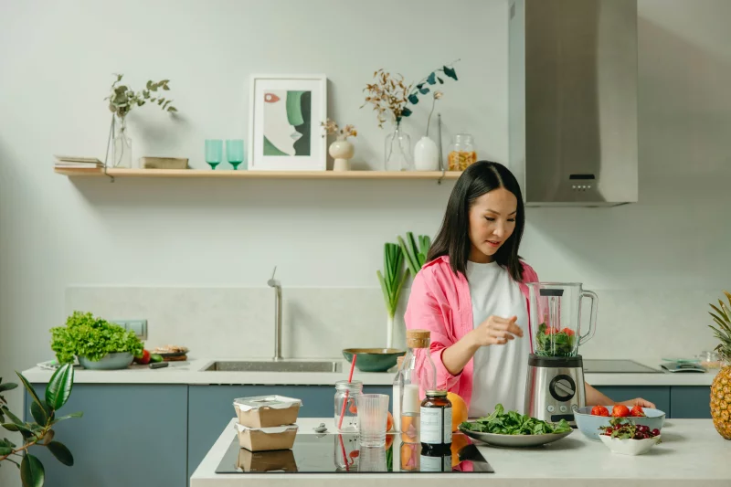 Asian woman blending fresh fruits and vegetables in a stylish kitchen setting.