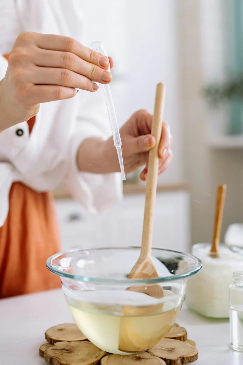 A detailed view of a person using a dropper and wooden ladle in a glass bowl for candle making.