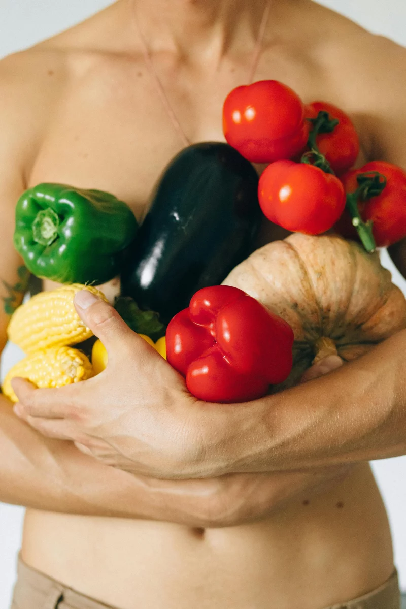 A man holding a variety of fresh vegetables including corn, pumpkin, peppers, tomatoes, and eggplant.