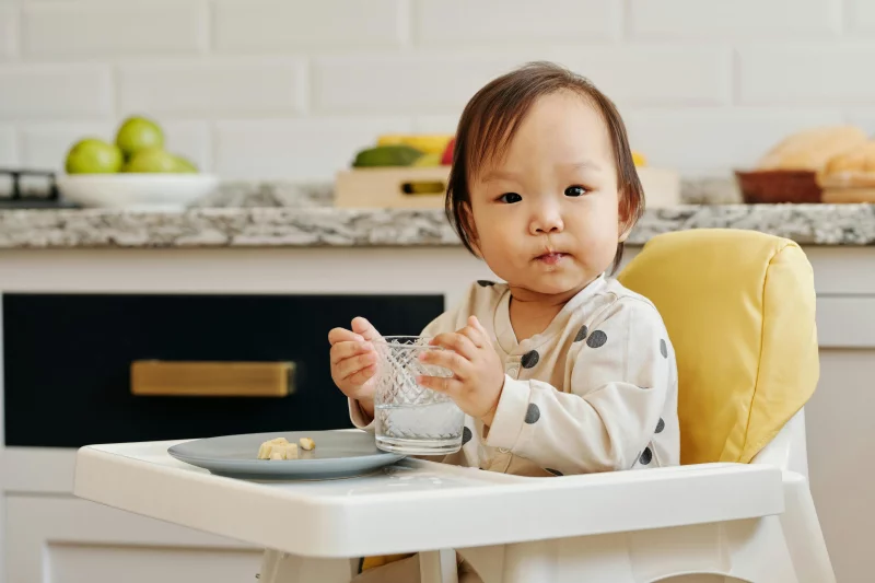 Adorable toddler sitting in a high chair, enjoying a meal and drink inside a cozy kitchen.