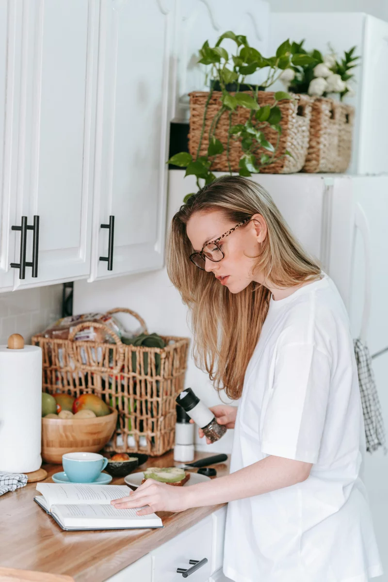 A woman multitasks in a kitchen, reading a book and preparing food on a countertop.